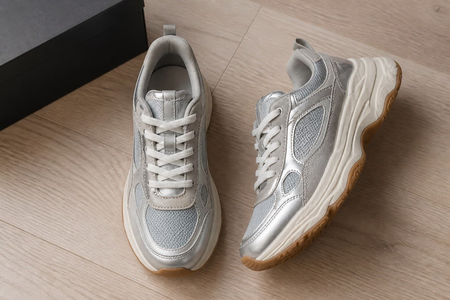 A pair of grey and cream sneakers on a wooden floor beside the corner of a black shoebox.