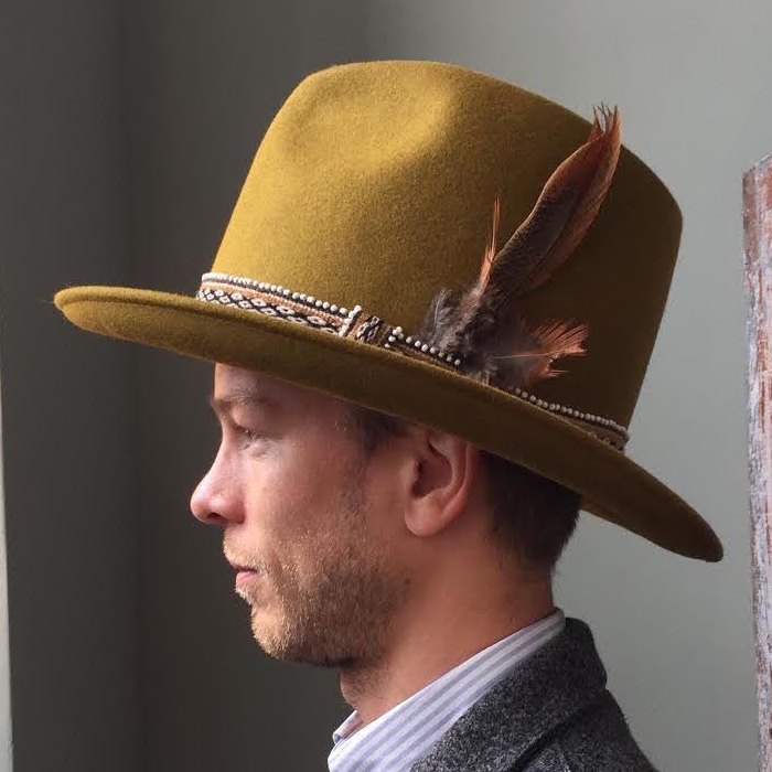 Johan Bjerke in profile, wearing a mustard-yellow felt hat with a feather in the band, a striped shirt and a grey tweed jacket